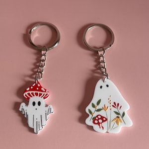 Può includere: Due portachiavi a forma di fantasma bianco con anelli e catene argentati. Un fantasma ha un fungo rosso e bianco sulla testa e l'altro ha un motivo floreale con fiori e funghi rossi e gialli.