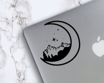 Puede incluir: Pegatina en blanco y negro de una luna creciente con montañas y estrellas en su interior. La pegatina está en un portátil plateado.