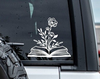 May include: White vinyl decal of an open book with flowers growing out of it. The decal is on the back window of a car.