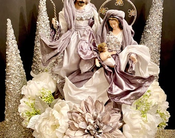 May include: A purple and white nativity scene centerpiece with a silver glitter Christmas tree on each side. The centerpiece features a white and purple fabric base with a white fabric flower and a purple flower.