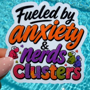 Puede incluir: Una pegatina de vinilo troquelada con el texto negro "Fueled by" sobre el texto naranja "anxiety &" y el texto morado "nerds" sobre la palabra "Clusters" con una textura de caramelo multicolor. Se incluyen pequeñas criaturas de dibujos animados rosas, verdes y moradas.