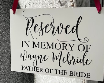 Könnte beinhalten: Ein weißes Schild mit schwarzem Text, auf dem steht "Reserviert in Gedenken an Wayne McBride Vater der Braut". Das Schild hängt an einem silbernen Stuhl mit einem roten Band.