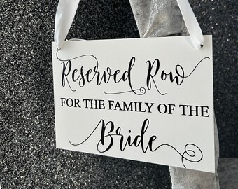 Könnte beinhalten: Ein weißes Schild mit schwarzem Text, der "Reservierte Reihe für die Familie der Braut" lautet. Das Schild hängt an einem grauen Samtband mit weißen Satinband-Bändern.