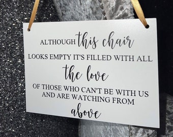 Könnte beinhalten: Ein weißes Schild mit schwarzem Text, der lautet: "ALTHOUGH this chair LOOKS EMPTY IT'S FILLED WITH ALL the love OF THOSE WHO CAN'T BE WITH US AND ARE WATCHING FROM above". Das Schild hängt an einem goldenen Band, das um einen grauen Samtstuhl gebunden ist.