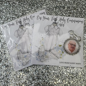 May include: Two white cards with a watercolour illustration of a boy in a white outfit. The cards say "On Your First Holy Communion" with the name "Jay" on one card and "Disin" on the other. A silver charm with a photo of a man is attached to the card with the name "Disin". The charm has the text "Love From Nanny Mary" on the back.
