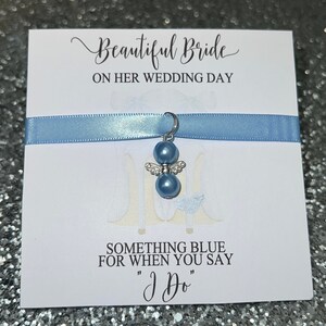以下が含まれることがあります： 青いリボンと銀の天使の羽根が付いた青いチャームが付いた白いカード。カードには「美しい花嫁、結婚式の日」と「あなたが「はい」と言うときに何か青いもの」と書かれています