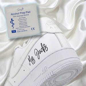 May include: A white Air Force 1 sneaker with the text "Ms. Jacks" in black script. An alcohol prep pad is in the background, with text including "70% Isopropyl Alcohol". The sneaker is on a white satin surface.