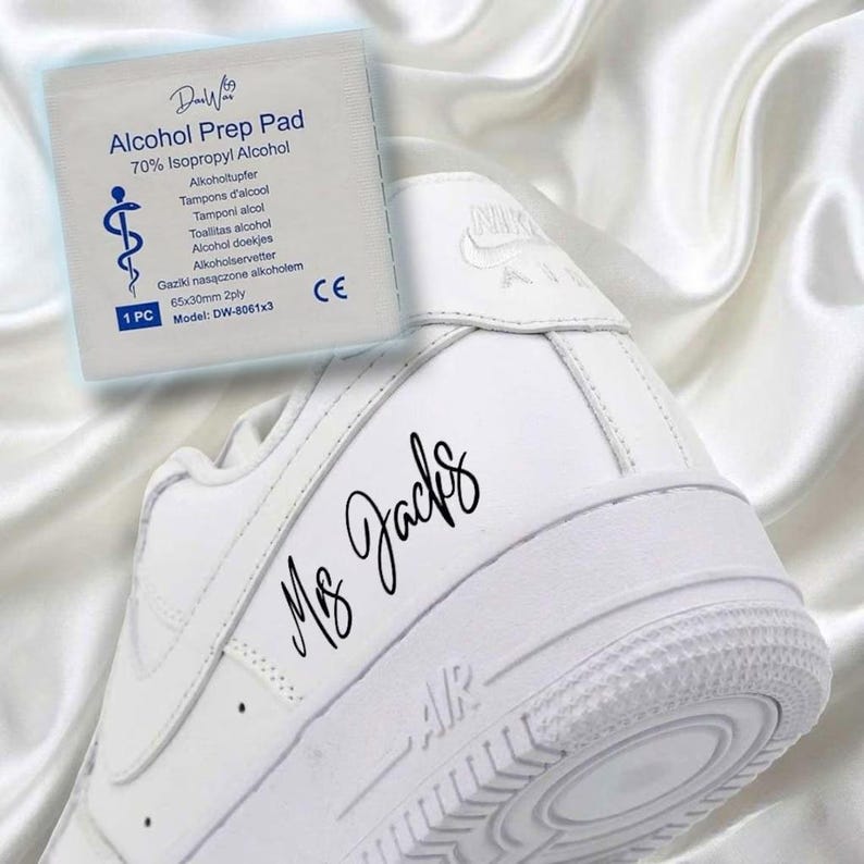 May include: A white Air Force 1 sneaker with the text "Ms. Jacks" in black script. An alcohol prep pad is in the background, with text including "70% Isopropyl Alcohol". The sneaker is on a white satin surface.
