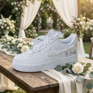 May include: White leather sneaker with pearl embellishments on the Nike swoosh and laces. The text "Ms. Jacks" is written on the side. The shoe is decorated with white flowers and greenery, suggesting a wedding theme.