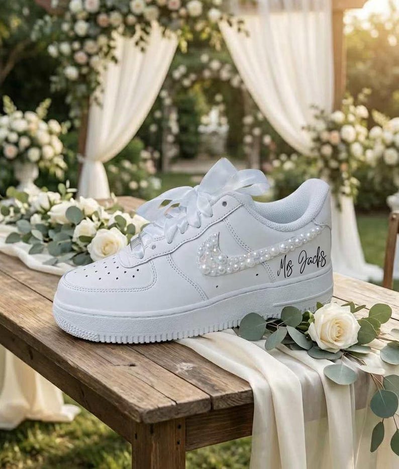 May include: White leather sneaker with pearl embellishments on the Nike swoosh and laces. The text "Ms. Jacks" is written on the side. The shoe is decorated with white flowers and greenery, suggesting a wedding theme.