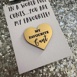 May include: A white card with the text "IN A WORLD FULL CUNTS... YOU ARE MY FAVOURITE!" and a gold heart-shaped pin that reads "MY FAVOURITE CUNT". The pin is attached to the card.
