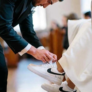 May include: White sneakers with black swoosh logos and the text "Mr. Jacks" are being laced up. The sneakers are on a person wearing a white dress. A person in a black suit is tying the laces.
