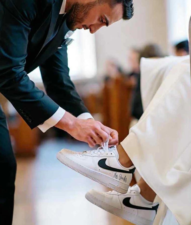 May include: White sneakers with black swoosh logos and the text "Mr. Jacks" are being laced up. The sneakers are on a person wearing a white dress. A person in a black suit is tying the laces.