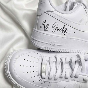 May include: Two white sneakers with laces, one atop the other. The top shoe has the text "Mrs Jacks" in black script. The bottom shoe has a metal lace tag. The shoes are on a white, textured surface.
