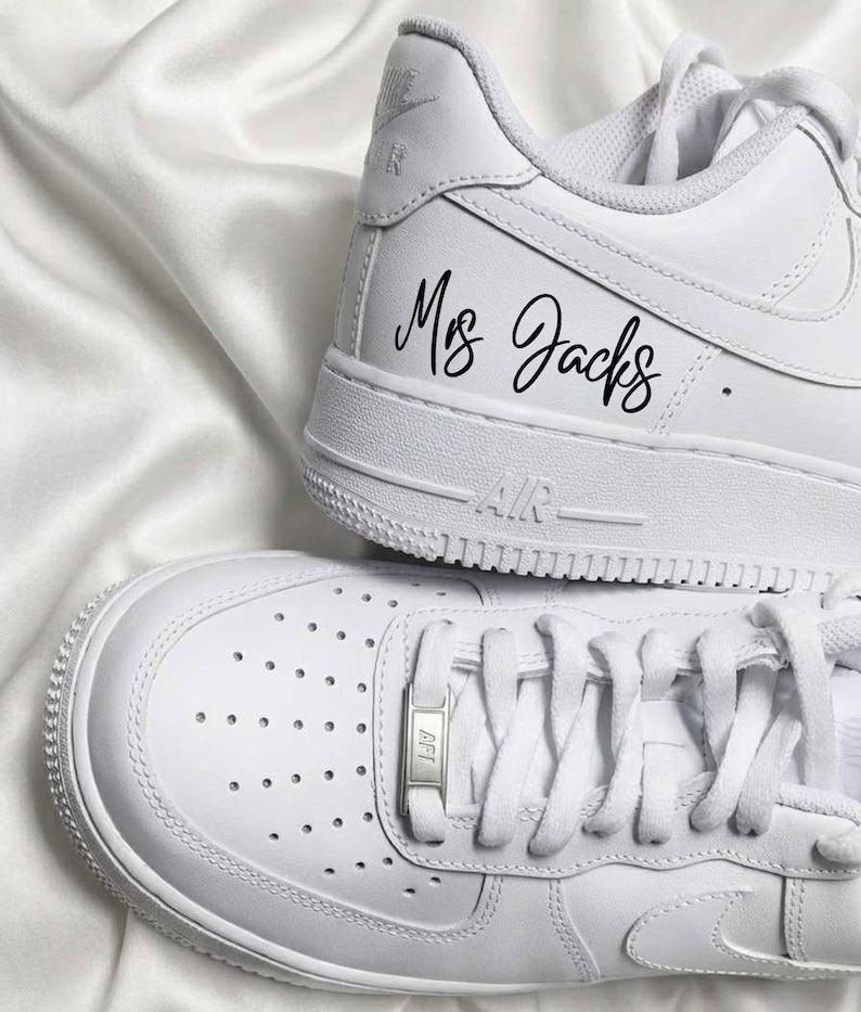May include: Two white sneakers with laces, one atop the other. The top shoe has the text "Mrs Jacks" in black script. The bottom shoe has a metal lace tag. The shoes are on a white, textured surface.