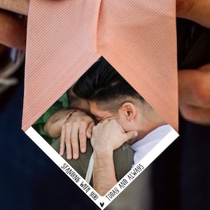 Puede incluir: Una corbata rosa con un inserto blanco en forma de diamante que presenta una foto de dos personas abrazándose. El texto "Standing with you" y "Today and always" está impreso en el inserto.