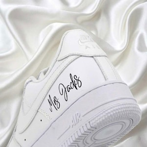 May include: A white Nike Air Force 1 sneaker with the text "Ms. Jacks" in black script on the side. The shoe is set against a white, silky background. The sole features a textured design.