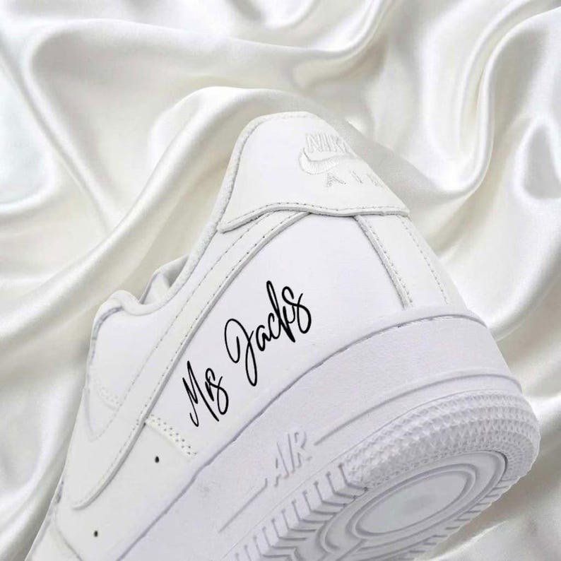 May include: A white Nike Air Force 1 sneaker with the text "Ms. Jacks" in black script on the side. The shoe is set against a white, silky background. The sole features a textured design.