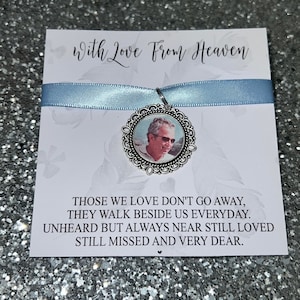 Op de afbeelding: Een witte kaart met een blauw lint en een zilveren bedel met een foto van een man. De kaart heeft de tekst "With Love From Heaven" en "Those we love don't go away, they walk beside us everyday. Unheard but always near still loved still missed and very dear."