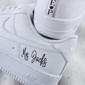 Può includere: Un paio di scarpe da ginnastica bianche con la scritta "AIR" sulla suola. Il testo "Ms Jacks" è stampato sul lato di una scarpa e le iniziali "E P" con un simbolo a cuore sono sul tallone dell'altra scarpa.