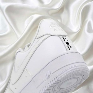 May include: White leather sneaker with the word "AIR" on the sole. The heel features the initials "E" and "P" with a heart symbol. The Nike logo is visible on the heel. The shoe is set against a white satin background.