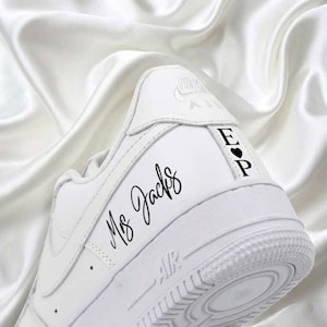 May include: White Nike Air Force 1 sneaker with custom black text. The text "Ms. Jacks" is written in cursive on the side. The heel features the initials "E" and "P" with a heart symbol. The shoe is set against a white satin background.