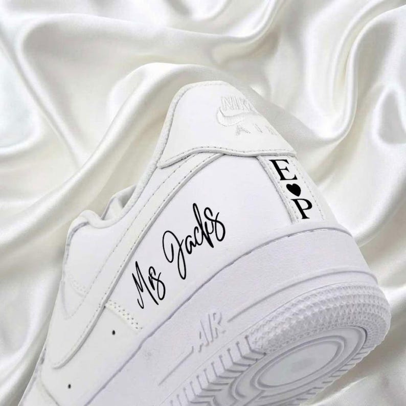 May include: White Nike Air Force 1 sneaker with custom black text. The text "Ms. Jacks" is written in cursive on the side. The heel features the initials "E" and "P" with a heart symbol. The shoe is set against a white satin background.
