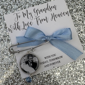 Peut inclure: Un ruban bleu noué autour d'une épingle de sûreté argentée avec un charme photo attaché. Le charme a une photo en noir et blanc d'un homme et d'une femme. Le texte sur la carte dit "To My Grandson With Love From Heaven" et "With You Today, Tomorrow And Forever".