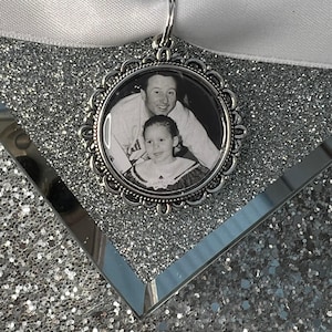 May include: A silver-toned photo charm with a black and white photo of a man and a young girl. The charm is attached to a white ribbon.