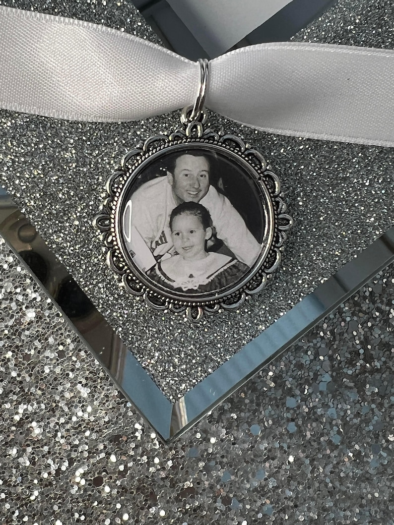 May include: A silver-toned photo charm with a black and white photo of a man and a young girl. The charm is attached to a white ribbon.
