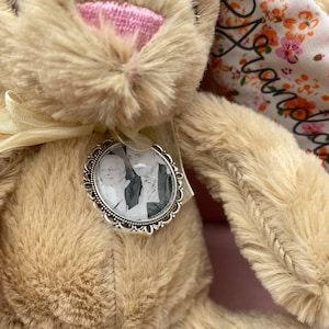 May include: A beige plush bunny with a pink nose and a silver locket containing a black and white photo. The bunny's ear is decorated with a floral fabric with the word "Grandma" embroidered on it.