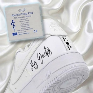 May include: White Nike Air Force 1 sneakers with custom black lettering that reads "Ms Jacks" on the side and initials "E P" with a heart on the heel. An alcohol prep pad is in the background.