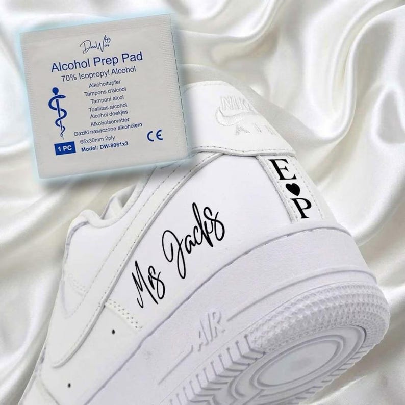 May include: White Nike Air Force 1 sneakers with custom black lettering that reads "Ms Jacks" on the side and initials "E P" with a heart on the heel. An alcohol prep pad is in the background.