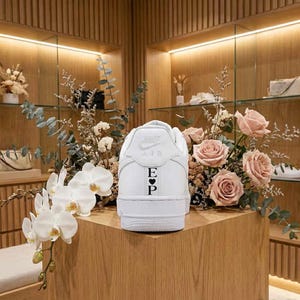 May include: A white sneaker with the Nike Air logo and the initials "E" and "P" with a heart symbol. The shoe is displayed on a wooden surface, surrounded by flowers and greenery.