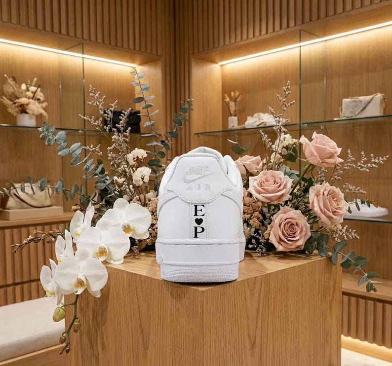 May include: A white sneaker with the Nike Air logo and the initials "E" and "P" with a heart symbol. The shoe is displayed on a wooden surface, surrounded by flowers and greenery.