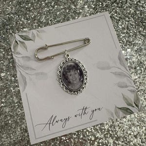 Op de afbeelding: Zilverkleurige herdenkingsspeld met een ovale fotohanger en decoratief frame. De speld is bevestigd aan een kaart met de tekst "Always with you" in script. De achtergrond is een sprankelend, glinsterend oppervlak.