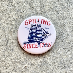 May include: A white button with red and blue text that reads "Spilling the Tea Since 1773". A blue and white illustration of a sailing ship is in the center of the button.
