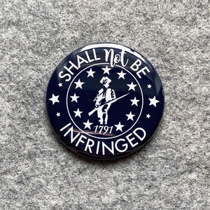 May include: A blue and white button with the text "Shall Not Be Infringed" and a silhouette of a person holding a rifle. The button also has a circle of stars around the text and the year "1791" in the center.