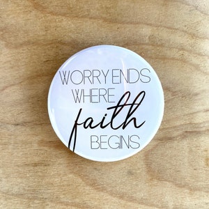 May include: White round button with black text that reads "Worry ends where faith begins".