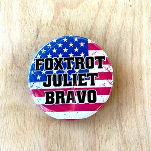 May include: A red, white, and blue button with a distressed American flag design. The text "FOXTROT JULIET BRAVO" is printed in black on the button.