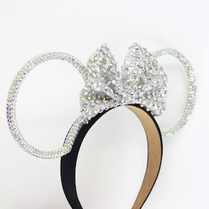May include: Silver sparkly mouse ears headband with a large bow. The headband is black with a brown backing. The ears are made of a silver sparkly material and are shaped like Mickey Mouse ears.