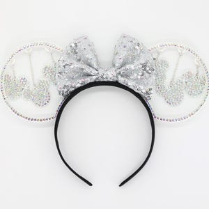 May include: A silver sequined bow and matching ears on a black headband. The ears are clear with a silver Mickey Mouse design. The headband is black and the overall design is sparkly and festive.