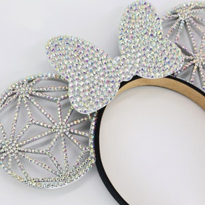 May include: A pair of silver sparkly mouse ears with a black headband. The ears are covered in iridescent rhinestones and have a geometric design.