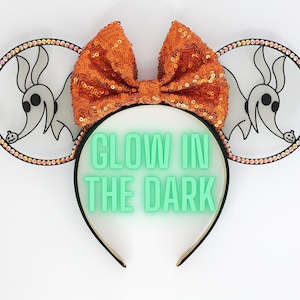 May include: A pair of white mouse ears with a glow-in-the-dark headband. The ears feature a black and white image of Zero, the ghost dog from The Nightmare Before Christmas, with a pumpkin in his mouth. The ears are decorated with iridescent rhinestones. The headband is adorned with a large orange sequined bow. The text "GLOW IN THE DARK" is printed in green on the headband.