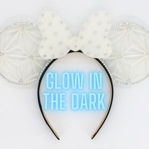 May include: White glow-in-the-dark Minnie Mouse ears with a white bow and a geometric design. The ears are decorated with rhinestones.