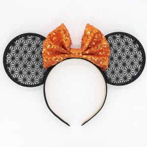 May include: A black headband with two black circular ears featuring a geometric pattern. An orange sequined bow is centered on the headband. The ears and bow are set against a white background.