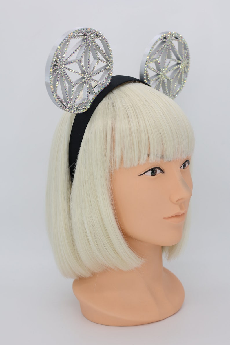 May include: A silver headband with two large, round, sparkly, geometric-shaped ears. The headband is black and the ears are decorated with iridescent rhinestones.