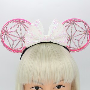May include: Pink mouse ears headband with a white sparkly bow. The ears are decorated with a geometric design and rhinestones.