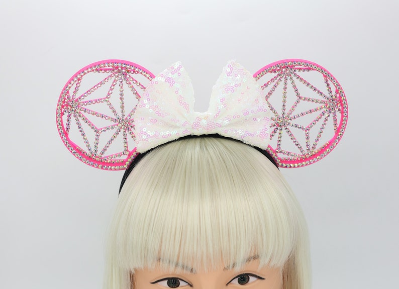 May include: Pink mouse ears headband with a white sparkly bow. The ears are decorated with a geometric design and rhinestones.