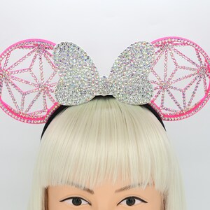 May include: Pink mouse ears headband with a large, sparkly, silver bow and a geometric design on the ears.